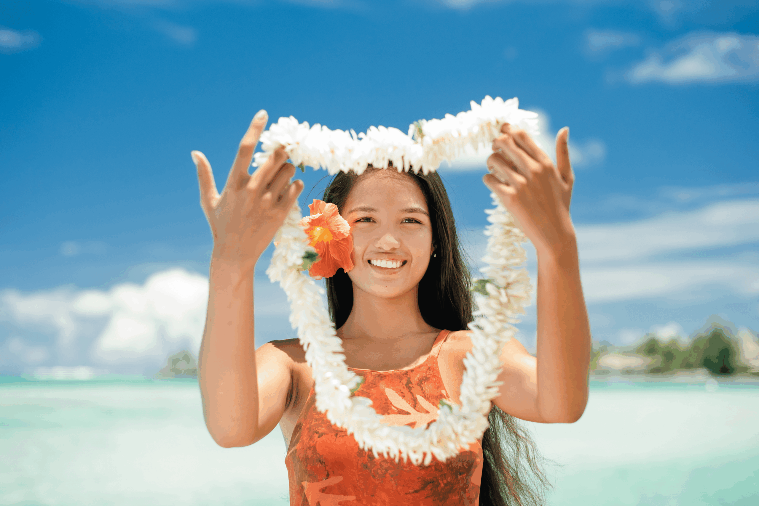 Jeune femme polynésienne souriante à Moorea, portant une fleur d'hibiscus à l'oreille et tenant un collier de fleurs de tiare traditionnel face au lagon turquoise