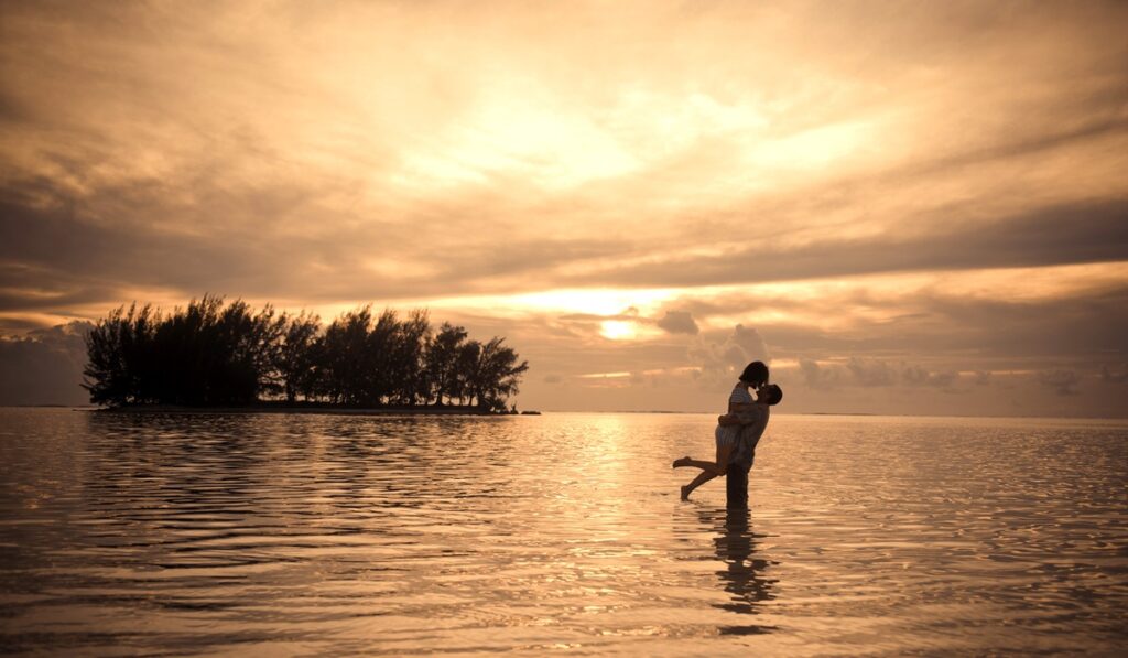 Couple enlacé dans l'eau au coucher du soleil face à une île polynésienne